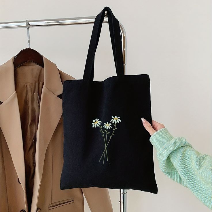 Black tote bag with daisy design held in front of beige coat on a rack.