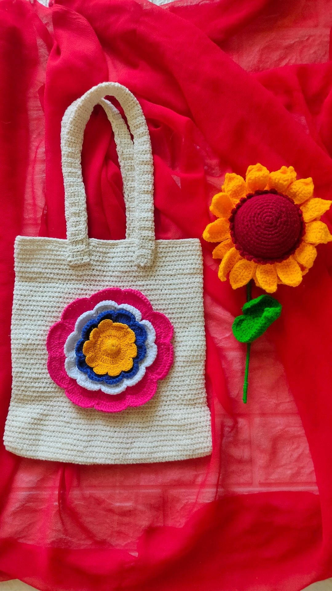 Crocheted white bag with colorful flower design on red fabric, accompanied by crocheted sunflower.