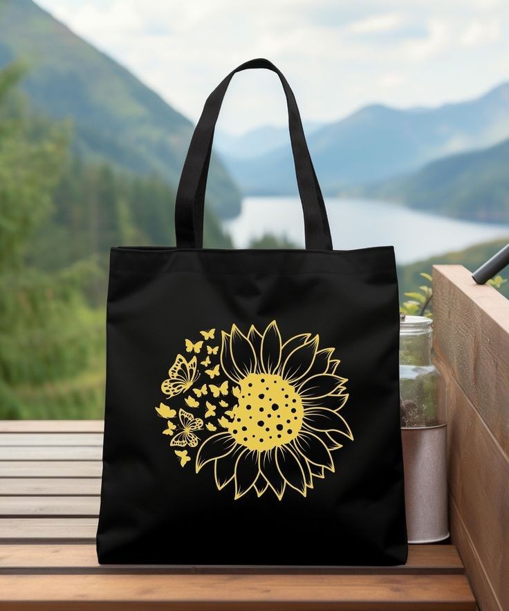 Black tote bag with yellow sunflower and butterfly design on a wooden table overlooking a scenic mountain view.