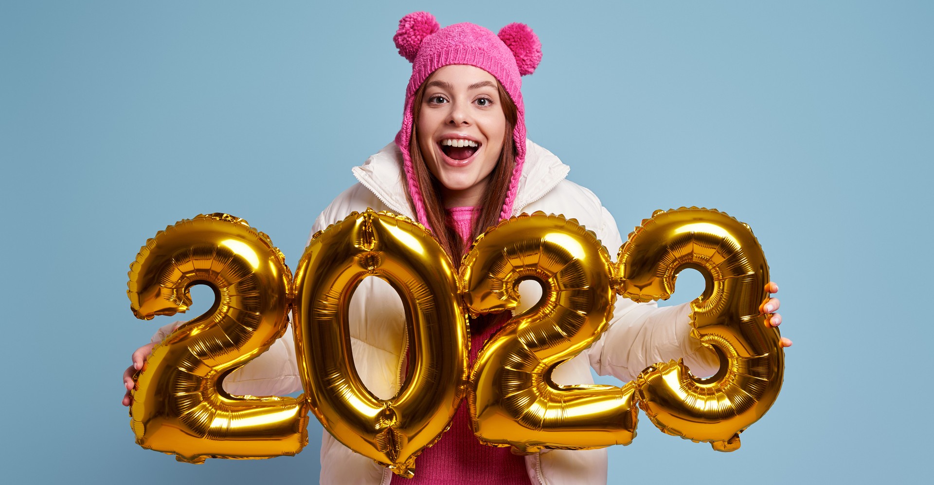 Excited young woman in winter coat holding gold colored numbers against blue background